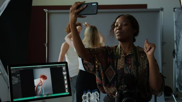 African American female photographer holding smartphone and filming vlog about shoot day in photo studio, stylist preparing model in background