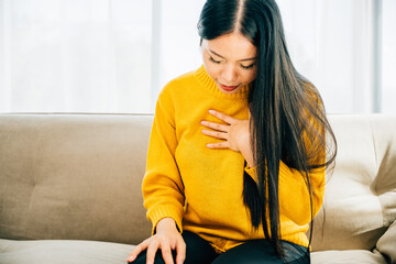 Illustrating heart attack, Asian woman on sofa holds her left chest in pain requiring immediate...