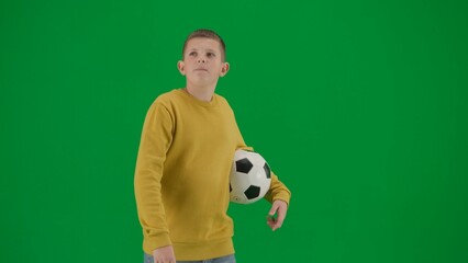 Fototapeta na wymiar Portrait of kid boy on chroma key green screen. Schoolboy in jeans walking holding football ball and looking around. Middle body side shot.