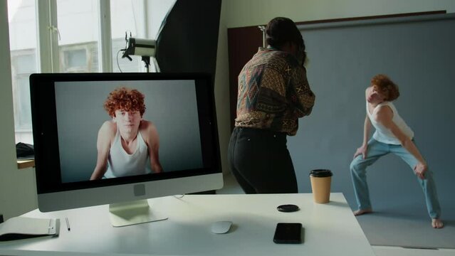 Female photographer taking pictures of young male model against gray cyc wall in studio, images appearing on computer monitor during tethered shooting
