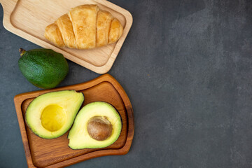 Top view of halved avocado on plate and croissant on a black background