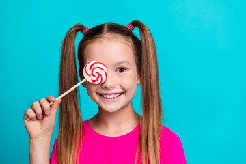 Portrait of toothy beaming nice small girl dressed pink t-shirt holding lollipop on eye smiling...