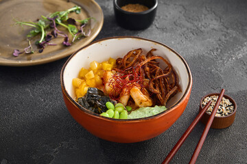 A vibrant shrimp and mango salad bowl, topped with crispy onions, edamame, and seaweed, served with chopsticks on a textured grey backdrop