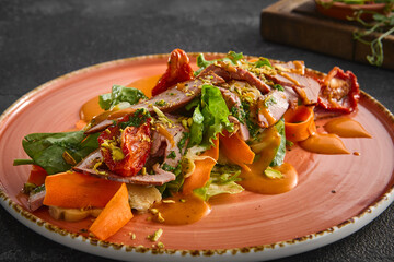A beautifully plated duck salad with vibrant carrot puree, served on a rustic peach-colored plate against a black textured backdrop