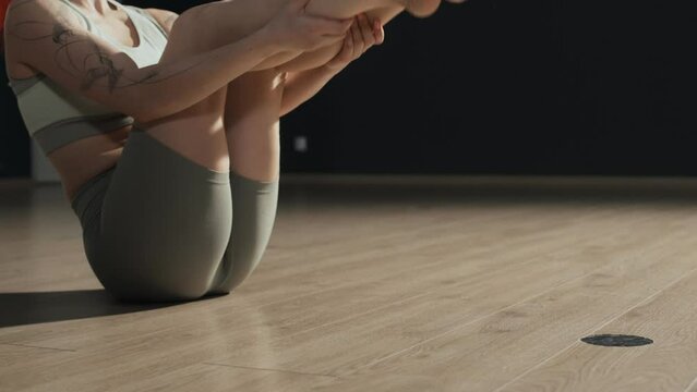 Tattooed red haired Caucasian young woman wearing tight activewear stretching her legs and back on parquet floor at minimalist sports studio with black walls