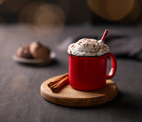 Red mug with hot chocolate or cocoa with whipped cream on a wooden stand with cinnamon sticks  on a dark background