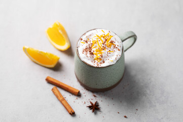 Mug of hot chocolate with whipped cream and orange zest on a light gray background with a cinnamon sticks  and fresh orange slices.