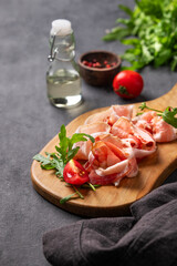 Snack wooden board with bacon or prosciutto on a dark background with spices, tomatoes and herbs.