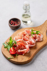 Snack wooden board with bacon or prosciutto on a light background with spices, tomatoes and herbs.