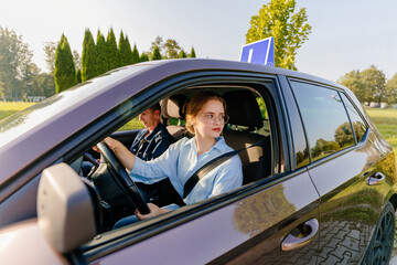 Teenage girl driving car with blue L plate on a roof. Instructor sitting next to woman trying to learn to park a vehicle at summer sunny day.