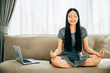 Asian woman meditates in lotus pose on sofa with laptop finding balance. Businesswoman embraces...