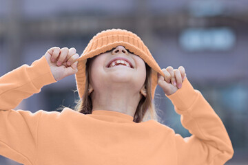 Trendy color of the 2024 year peach fuzz. Cute little caucasian girl eight years old with blonde hair smiling outdoor. Kid wearing stylish shirt and knitted hat