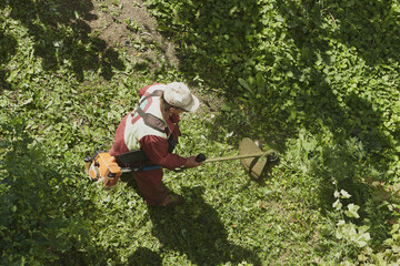 The gardener in overall and safety helmet trims overgrown grass by grass cutter