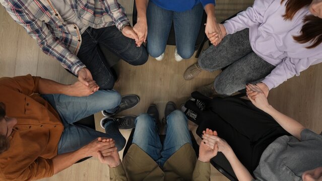 In the frame above sits on a chair in the center, middle aged man, psychologist, mentor. Opposite sits his group they have a meeting, therapy. They are holding hands and talking, communicating