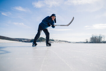 An elderly man practices stricking the puck with hockey stick on a frozen lake in winter. - obrazy, fototapety, plakaty