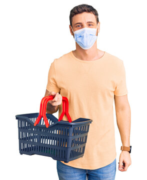 Handsome young man with bear wearing shopping basket and medical mask looking positive and happy standing and smiling with a confident smile showing teeth