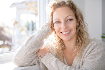 Happy attractive blonde woman at home close to a window - obrazy, fototapety, plakaty