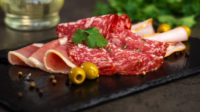Close up of fresh slices cold cuts, sausage ham bacon jamon on stone cutting board with olives and parsley. Meat pork beef. Food