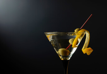 Glass of classic dry martini cocktail with green olives and lemon peel on a black background.