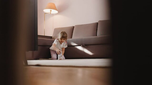 A cute little girl in pajamas sits on the floor at home near the sofa and plays with a toy cat. The mechanical cat is moving towards the child, and she is trying to catch it.