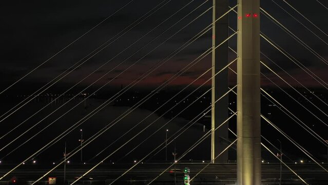 A cable-stayed highway bridge with tall concrete pylons illuminated by architectural lighting at night. Vertical climb up along the pylons against the background of night city lights with camera