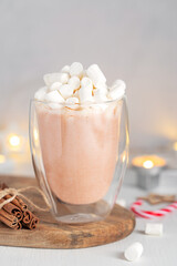 Homemade sweet cocoa drink or hot chocolate decorated with topping of marshmallow sweets served in transparent glass cup or mug with cinnamon sticks on wooden board on white table against candle light