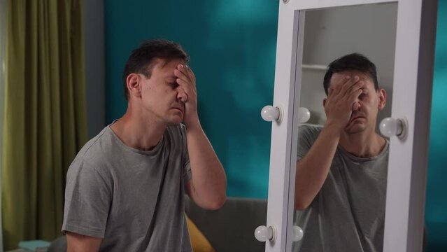An exhausted, tired man suffering from a mental disorder looks at his reflection in a large mirror close up. A man with bipolar disorder follows the movement of his hand, hides his face in his palms.