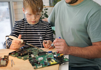Father with his son spend time together. Close-up of a man teaching his boy at home to solder computer spare parts. Education moment during parenthood. Togetherness concept.
