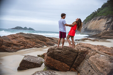 Casal jovem sorridente, se divertindo, conversando, em pe sobre uma pedra na praia. - obrazy, fototapety, plakaty
