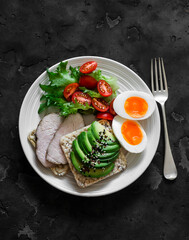 Healthy delicious breakfast on a dark background - sandwich with avocado and baked turkey, boiled egg, fresh vegetable salad with cherry tomatoes