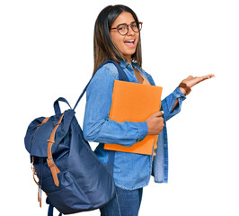 Young latin girl wearing student backpack and holding books pointing aside with hands open palms...
