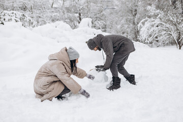 Fototapeta na wymiar Young couple having fun making building snowman in winter snowy season in city park covered hill, holiday vacation weekend, enjoying spending time together 