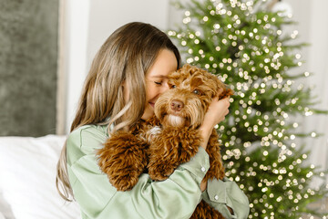 A Beautiful young woman with a golden brown labradoodle dog on christmas tree , bokeh light garland bacground. Cute Family puppy and girl play at home, new year decorated interior at Christmas 