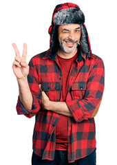 Middle age hispanic man with grey hair wearing fluffy earmuff hat smiling with happy face winking at the camera doing victory sign. number two.