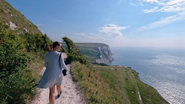 Female tourist walks with camera along picturesque coastal footpath above high cliffs. Young holidaymaker is exploring beautiful White Cliffs of Dover that stretch above deep blue ocean on a sunny day