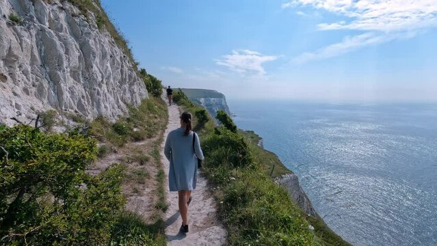 Following a female photographer exploring along beautiful White Cliffs of Dover. Young woman is walking on a scenic coastal footpath that stretches on the edges of a high cliff above deep blue ocean.