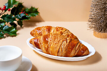 Christmas breakfast. Fresh delicious croissants lying on plate on beige background. Next to it is small shiny decorative Christmas tree, coffee cup and branch of holly