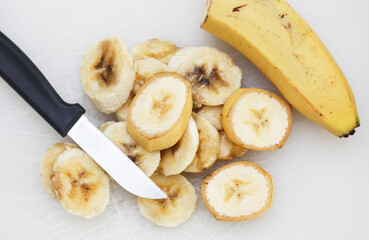 Bananas on white cutting board. Ripe banana slices isolated on white. Sunlight fruits. Quick healthy morning snack. Yellow fruits with brown stains. Cut banana slices. Small kitchen knife.