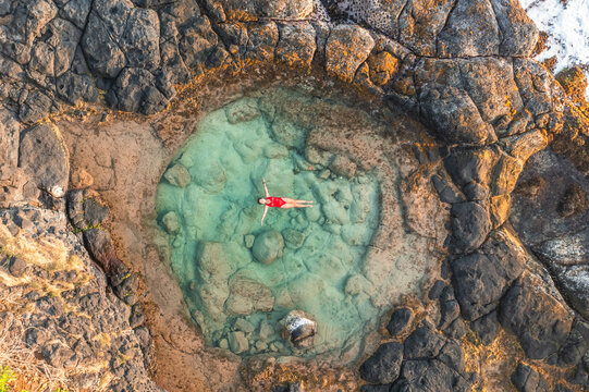 Aerial view of a woman swimming in a natural rock pool along the coastline with waves breaking on the cliffs near Gris Gris beach, Savanne, Mauritius.