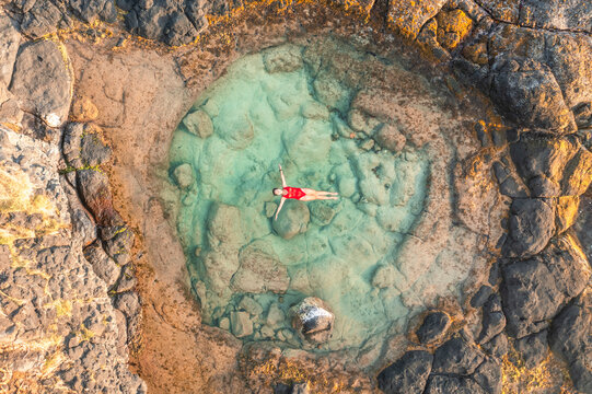 Aerial view of a woman swimming in a natural rock pool along the coastline with waves breaking on the cliffs near Gris Gris beach, Savanne, Mauritius.