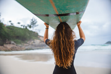 Young woman on the beach holding her surfboard above her head. Rear view.