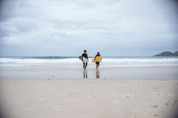 Couple of surfers walking along the beach with their boards under their arms, talking.