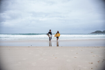 Couple of surfers walking along the beach with their boards under their arms, talking.