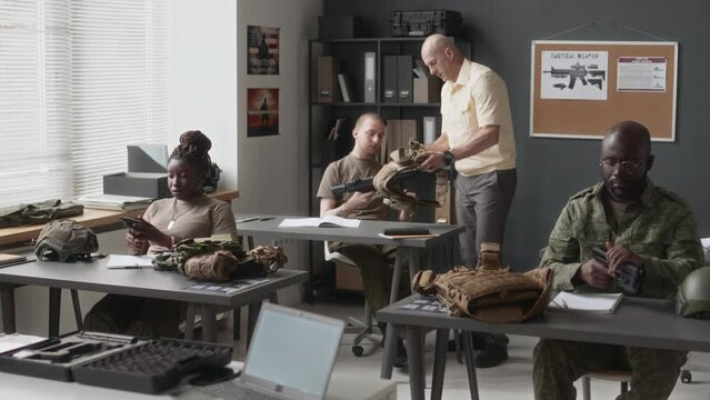 Full shot of Caucasian military instructor walking around classroom and working with diverse cadets who learning how to use firearm and plate carriers during training class in service academy