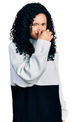 Young hispanic woman with curly hair wearing casual sweater smelling something stinky and disgusting, intolerable smell, holding breath with fingers on nose. bad smell