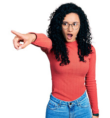 Young hispanic woman with curly hair wearing glasses pointing with finger surprised ahead, open mouth amazed expression, something on the front