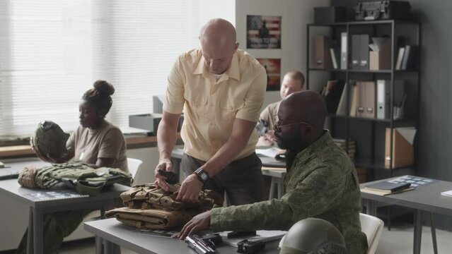 Side shot of Caucasian military instructor helping African American cadet with plate carrier and then watching him assembling hand gun during training class in academy