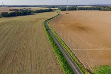 Rozległa równia pokryta porośniętymi trawą łąkami, lasami i brązowymi, zaoranymi polami uprawnymi. W centrum widać wąską asfaltową drogę. Zdjęcie wykonano z użyciem drona.