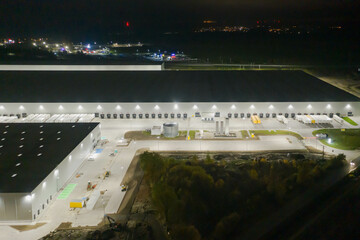 Centrum logistyczne w nocnych ciemnościach. Teren wokół jak i budynki oświetlone są sztucznym światłem elektrycznym. Zdjęcie zrobione przy użyciu drona.