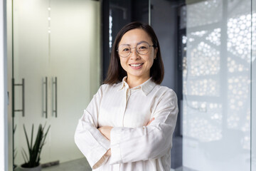 Portrait of young mature asian businesswoman entrepreneur, woman with crossed arms smiling and looking at camera, working inside office at workplace, standing near window in light clothes.